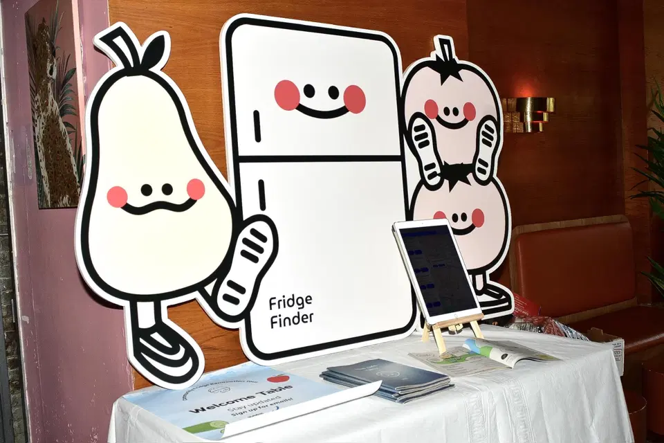 A table  on top of which stands a cardboard cutout of the Fridge Finder mascots. On top of the table are printed booklets, a welcome poster, and an iPad on a display stand.